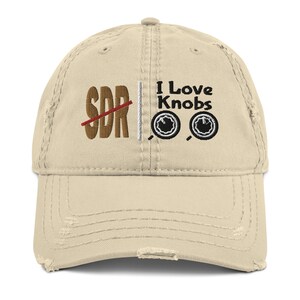 Peut inclure: Une casquette de baseball beige usée avec le texte "SDR" barré et le texte "I Love Knobs" avec deux illustrations de boutons noirs et blancs.