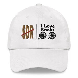 Peut inclure: Casquette de baseball blanche avec une broderie brune qui dit "SDR" avec une ligne rouge qui la traverse. Le côté droit de la casquette a une broderie noire qui dit "J'aime les boutons" avec deux boutons noirs et blancs.