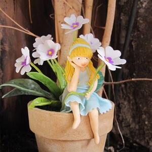 以下が含まれることがあります： 金髪の小さな磁器製のドールが、テラコッタ製の植木鉢の縁に座っています。ドールは薄い青色のドレスを着ており、手に花を持っています。鉢は、紫色の芯を持つ白い花で囲まれています。