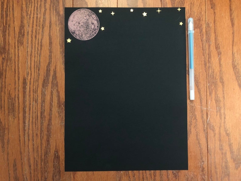 Puede incluir: Una hoja de papel negra con una luna rosa y estrellas doradas en la parte superior. Un bol&iacute;grafo azul est&aacute; en el lado derecho del papel.