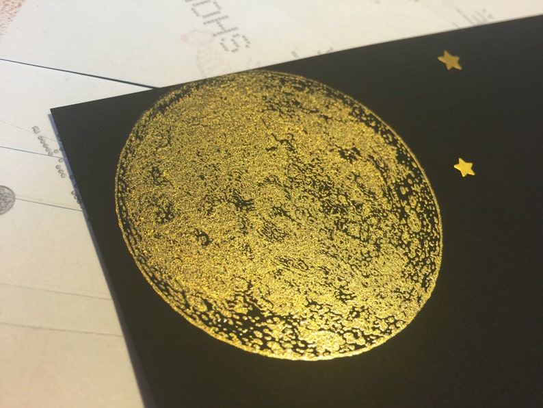 Puede incluir: Una tarjeta negra con una luna de l&aacute;mina dorada y dos estrellas de l&aacute;mina dorada. La luna tiene una textura de superficie detallada.