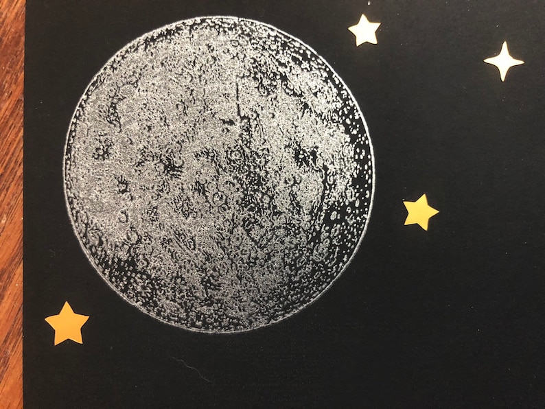 Puede incluir: Una luna plateada con una textura de superficie detallada sobre un fondo negro con cuatro estrellas doradas.