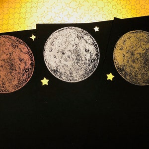 Puede incluir: Tres tarjetas negras con un dise&ntilde;o de luna dorada, plateada y cobre en cada una. Las lunas est&aacute;n impresas sobre un fondo negro con estrellas doradas.