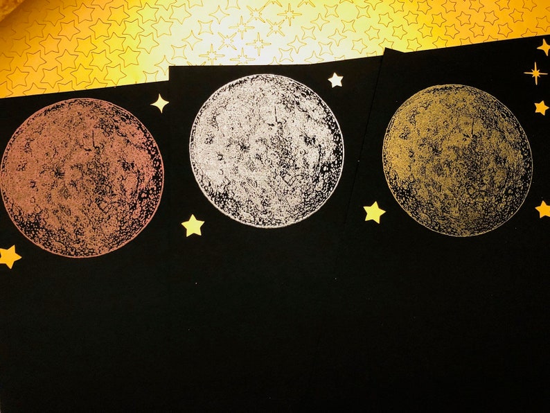 Puede incluir: Tres tarjetas negras con un dise&ntilde;o de luna dorada, plateada y cobre en cada una. Las lunas est&aacute;n impresas sobre un fondo negro con estrellas doradas.