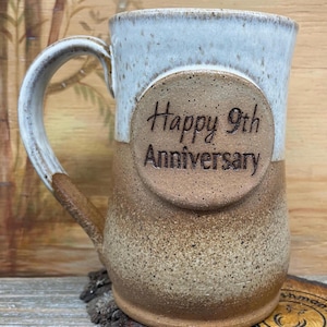 Könnte beinhalten: Eine braune und weiße Keramiktasse mit einem braunen Kreis auf der Seite, auf dem "Happy 9th Anniversary" steht.