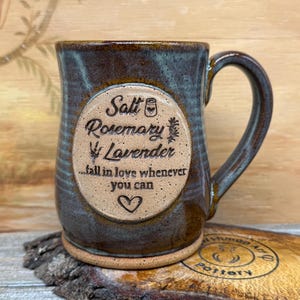 May include: A ceramic mug with a mottled blue and brown glaze featuring a central circular medallion. The text reads Salt, Rosemary, Lavender, ...fall in love whenever you can, with icons of a salt shaker, a sprig, and a heart.