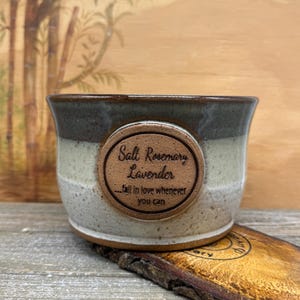 May include: A handcrafted ceramic bowl with a brown rim, a light green middle, and a speckled white base. A brown, oval-shaped tag is attached to the bowl, with the words "Salt Rosemary Lavender" and "...fall in love whenever you can."