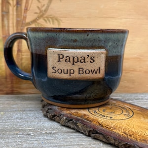 May include: A handmade ceramic soup bowl with a dark blue and brown glaze. The text "Papa's Soup Bowl" is displayed on a rectangular plaque attached to the bowl. The bowl sits on a wooden coaster.