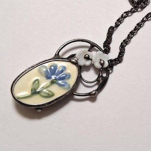 May include: A silver-toned necklace featuring an oval pendant with a blue flower design on a cream background. Two small white flower accents and a dark chain complete the necklace. The floral pendant is approximately 1.5 inches long.