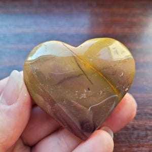 May include: A polished heart-shaped stone featuring a smooth, glossy surface with earthy tones of mustard yellow, brown, and deep maroon. The stone displays natural linear inclusions and dark veining patterns across its curved, symmetrical form.