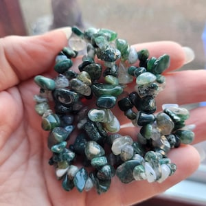 May include: A bracelet composed of small, polished green and white gemstones. The stones range in colour, from dark forest green to pale translucent white. The bracelet is held in a hand, highlighting the natural variations and textures of the stones.