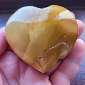 May include: A polished heart-shaped stone in shades of mustard yellow and golden brown. The surface is smooth and reflective with subtle natural veining and a small, dark circular indentation near the bottom point of the heart shape.