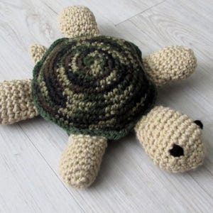 May include: A handmade crochet turtle toy. The turtle has a camouflage-patterned shell in shades of green, brown, and tan. The head, feet, and tail are a solid beige color. The turtle has small black eyes.
