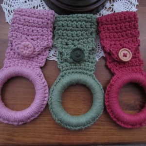 May include: Three handmade crochet towel holders in shades of pink, green, and red. Each holder features a loop for the towel and a button closure. The crochet work is visible, showcasing the texture and craftsmanship.