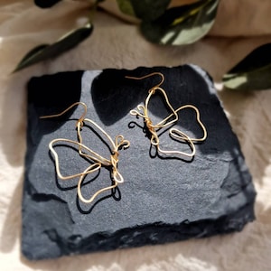 May include: A pair of gold-toned butterfly earrings made of wire. The earrings are delicate and have a simple design.