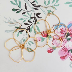 May include: A pair of gold-colored wire flower earrings. The earrings are crafted in the shape of open flowers with five petals. The earrings are displayed on a floral patterned background with pink and green accents.