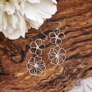 Könnte beinhalten: Silberdraht-Blumenohrringe mit dreidimensionalem Design. Die Ohrringe weisen ein mehrschichtiges Blumenmuster auf, wobei jede Blume aus zarten Drahtblütenblättern besteht. Die Ohrringe werden auf einer Holzoberfläche präsentiert.