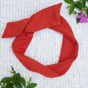 May include: A terracotta-colored fabric headband tied in a knot. The headband is on a white textured surface.
