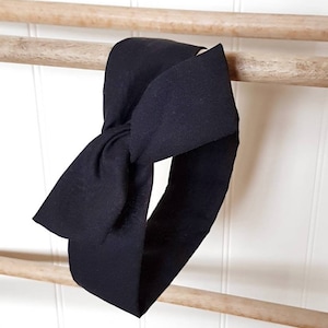May include: Black fabric headband tied in a bow. The headband is hanging on a wooden rack.