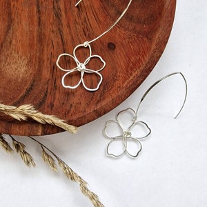 May include: A pair of silver-colored flower-shaped earrings. Each earring features a delicate, openwork floral design suspended from a curved wire. The earrings are displayed on a wooden surface with a sprig of dried grass.