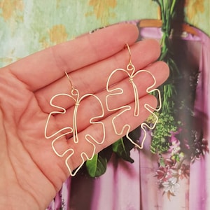 May include: Gold-coloured wire earrings shaped like monstera leaves. The earrings are displayed against a floral backdrop. The earrings have a hook closure.