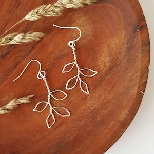 May include: A pair of silver wire earrings shaped like delicate branches with leaves. The earrings are displayed on a wooden surface.