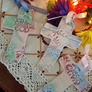 Puede incluir: Tres cruces de cerámica blanca con diseños florales y mensajes inspiradores. Las cruces están decoradas con colores pastel y tienen cintas atadas. Los mensajes en las cruces son "Nada es imposible con Dios", "gracia" y "Jesús vive".