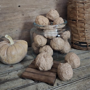 May include: A clear glass jar filled with round, textured, light brown objects, accompanied by a pale tan pumpkin with a green stem. A small wooden pallet sits in front of the jar, holding one of the round objects, with two more placed nearby.