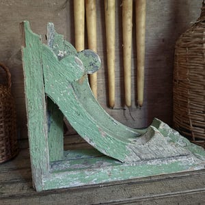 May include: A weathered green wooden architectural corbel featuring a curved, scroll-like design with visible paint distress and chipping. The structural bracket displays a flat base and a vertical back piece, showcasing a worn, aged texture.