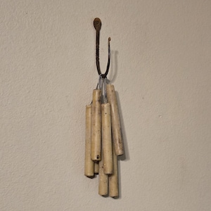 May include: A decorative wall hook with a dark, textured metal design. The hook holds a bundle of cylindrical, off-white ceramic pieces. The ceramic pieces have a speckled appearance and are arranged in a cascading pattern, adding a rustic touch to the wall decor.