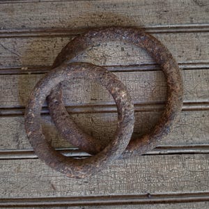 May include: Two circular metal rings with a heavily rusted, textured brown surface. The rings are linked together, with one ring partially overlapping the other, showcasing a weathered, oxidized appearance on their thick, rounded metal frames.