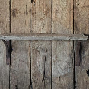 May include: A wall-mounted shelf featuring a rectangular, weathered gray wooden board supported by two dark, rusted metal L-shaped brackets. The shelf is attached to a vertical surface made of rough, unfinished vertical wooden planks.