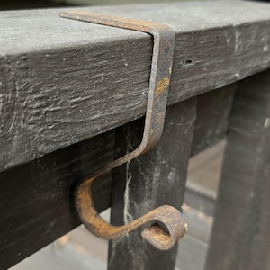 May include: A rusty metal hook, designed to hang over a ledge. The hook is a dark, aged metal with a curved bottom and a straight top section. The hook is attached to a dark wooden surface.