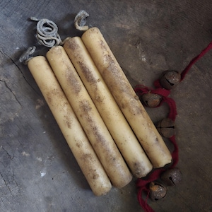 May include: Four cream-colored, rustic-style candles with brown speckling are arranged diagonally. The wicks are visible at the top. A strand of small, rusty bells on a red string is on the right. The candles rest on a weathered, wooden surface.