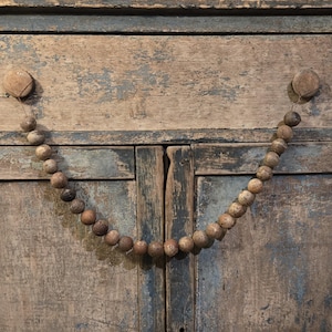 May include: A rustic wooden cabinet with a distressed blue and brown finish. A strand of round, textured wooden beads hangs between two wooden knobs, creating a decorative handle. The cabinet doors are closed.