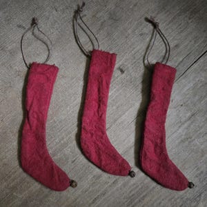 May include: Three red felt stockings with attached loops for hanging. Each stocking has a small bell at the toe. The stockings are displayed on a wooden surface.