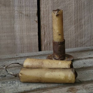 May include: Three vintage candles, two are lying on a wooden surface and one is in a rusty metal candle holder. The candles are a pale yellow color and have a rough texture.