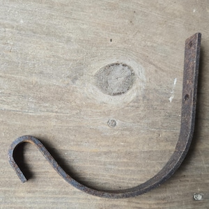 May include: A rustic, curved metal hook with a weathered, brown finish. The hook has a rectangular mounting plate with two holes for attachment and a curved lower section. The hook is designed for hanging items.