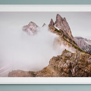 Puede incluir: Una fotografía de una cordillera con un pico cubierto de nubes. La montaña es rocosa y marrón con algunas manchas verdes de vegetación. Hay algunas personas de pie en la cima.