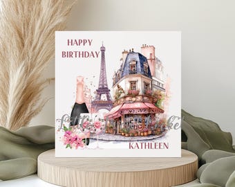 May include: A square birthday card featuring a watercolor illustration of Paris with the Eiffel Tower, a cafe, and a champagne bottle. The words "Happy Birthday" and "Kathleen" are printed on the card. The card is displayed on a wooden stand.