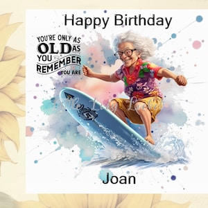 Puede incluir: Tarjeta de feliz cumpleaños con un fondo de acuarela que presenta a una mujer con cabello gris montando una tabla de surf. El texto en la tarjeta dice "You're only as old as you remember you are" y "Joan".