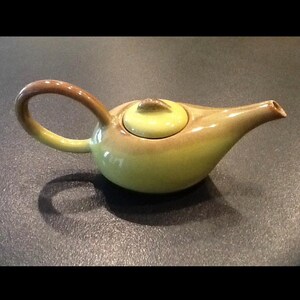 May include: A ceramic teapot featuring a rounded body with a light green glaze that transitions into a warm brown tone along the handle, spout, and lid. The teapot has a curved handle, a tapered spout, and a small, circular lid with a knob.
