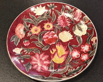 May include: A small, round, red plate with a gold rim. The plate is decorated with a floral pattern in shades of pink, yellow, and green.