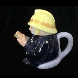 Puede incluir: Tetera de cer&aacute;mica con forma de bombero. La tetera presenta un casco amarillo, una chaqueta azul marino y un asa gris claro. El pico est&aacute; dise&ntilde;ado para parecerse a una manguera. Art&iacute;culo decorativo.