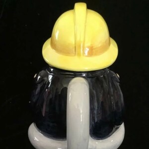 Puede incluir: Una tetera de cer&aacute;mica con forma de bombero. La tetera tiene un casco amarillo, un abrigo azul oscuro y un asa blanca. La tetera est&aacute; dise&ntilde;ada para parecerse a un uniforme de bombero.