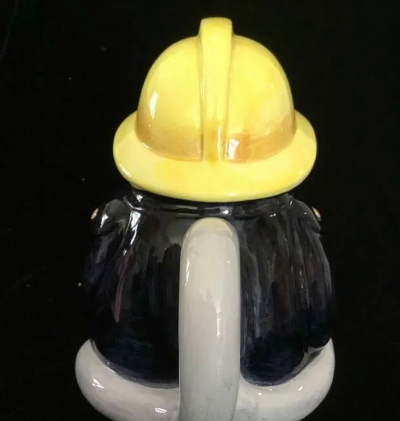 Puede incluir: Una tetera de cer&aacute;mica con forma de bombero. La tetera tiene un casco amarillo, un abrigo azul oscuro y un asa blanca. La tetera est&aacute; dise&ntilde;ada para parecerse a un uniforme de bombero.
