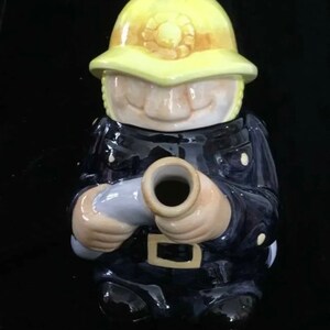 Puede incluir: Una figura de cer&aacute;mica de un bombero con uniforme azul marino y casco amarillo. La figura sostiene una manguera gris y tiene una cara sonriente. El casco tiene un emblema decorativo.