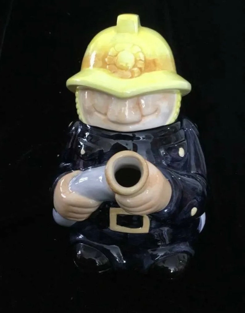 Puede incluir: Una figura de cer&aacute;mica de un bombero con uniforme azul marino y casco amarillo. La figura sostiene una manguera gris y tiene una cara sonriente. El casco tiene un emblema decorativo.