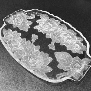 May include: An oval clear glass serving tray featuring a frosted, raised relief design of multiple blooming roses and leaves. The edges of the tray are scalloped, and the floral pattern is distributed across the surface of the glass.