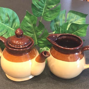 May include: A ceramic teapot and creamer set. The teapot has a lid and spout, and the creamer has a handle and spout. Both pieces are brown and tan with a glossy finish. Green leaves are in the background.